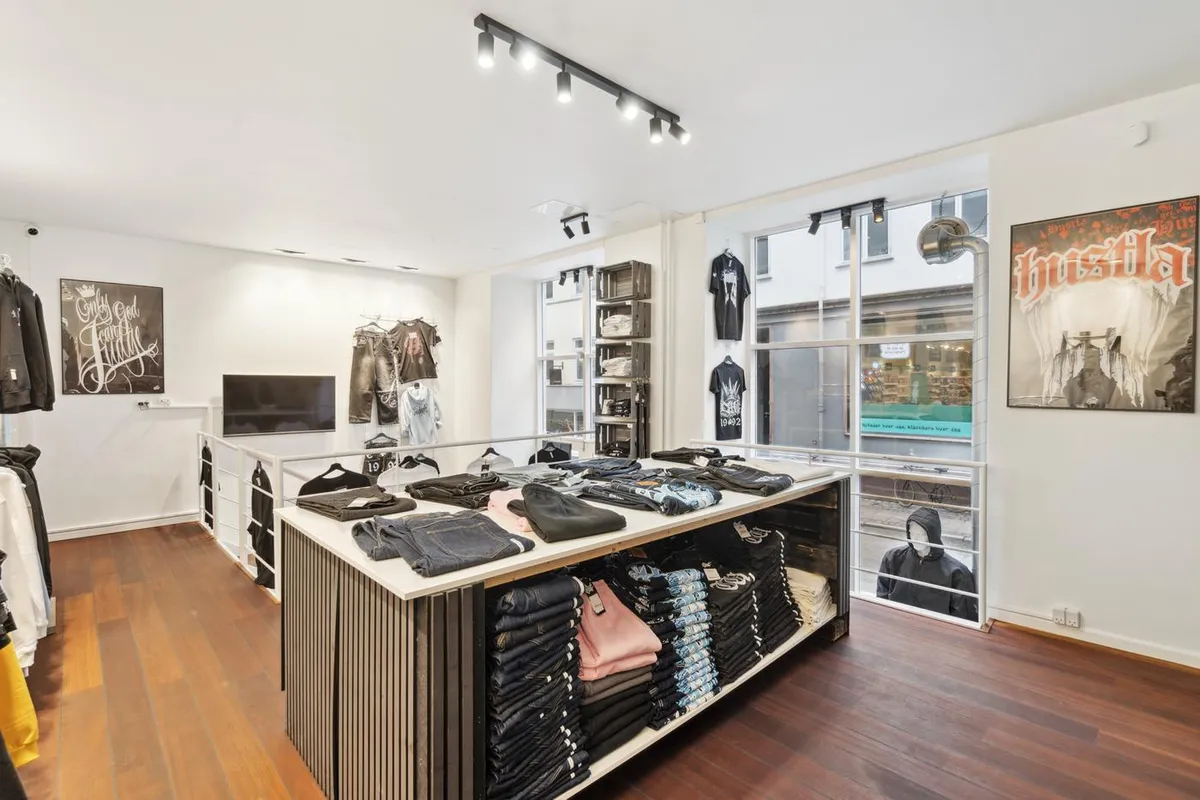 Indbydende butik med tøj på lager, mærket Bushila. Tøjet er udsat på snedkerborde og vægskabe. Plakater hænger på væggen.