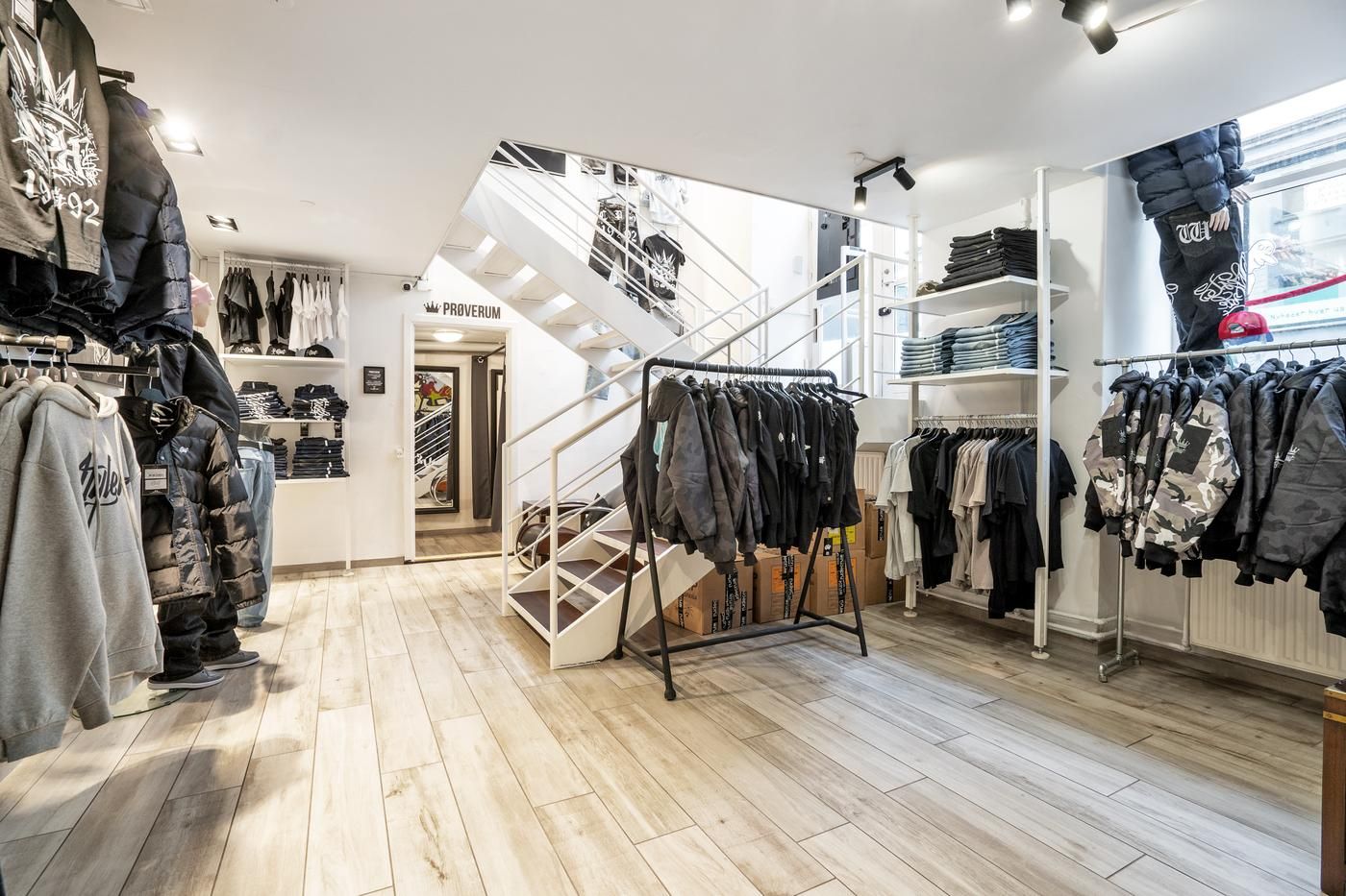 Hængende tøj på hylder og stående tøj på væggen. Støvler, jakker og t-shirts med forskellige mønstre og farver. En stige med