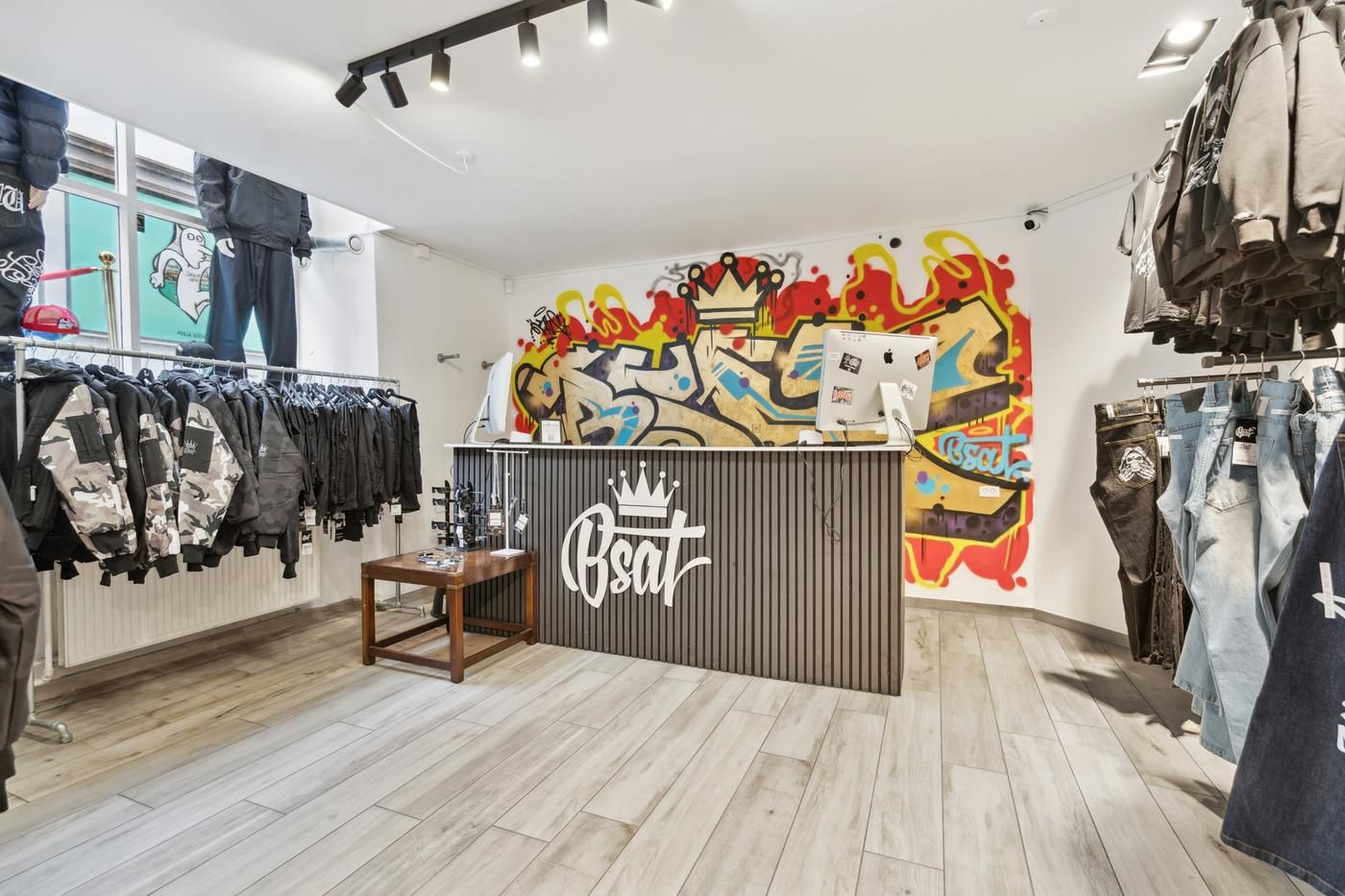 Butik med graffiti på væggen og tøj på hængere. Butiksfront med logo og skrift. To skærme og et bord på butiksfronten. Tøj