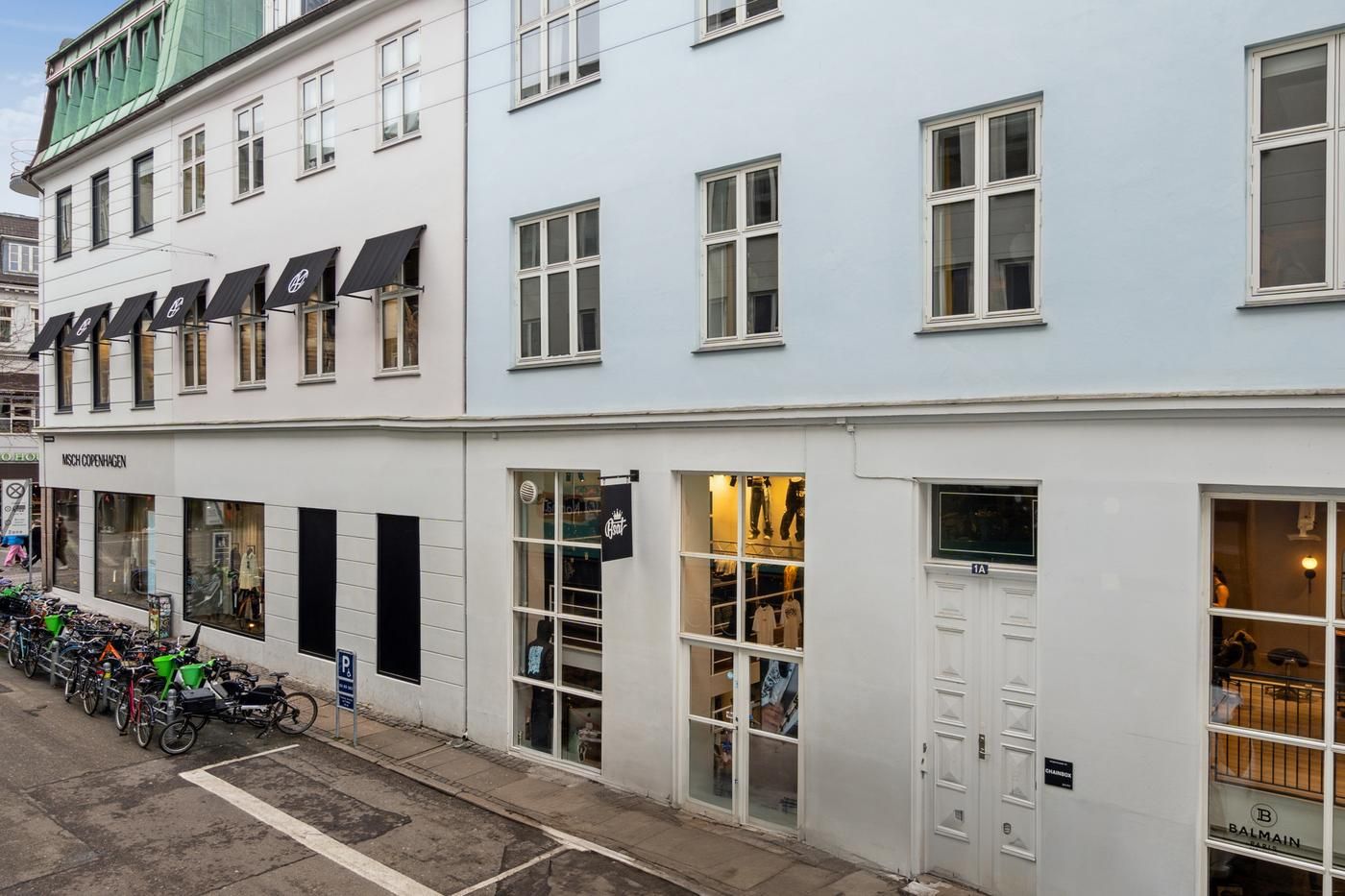 Butiksfront med lyse vinduer og sort banner med tekst og logo, butikker med navne som Misch Copenhagen og Balmain.