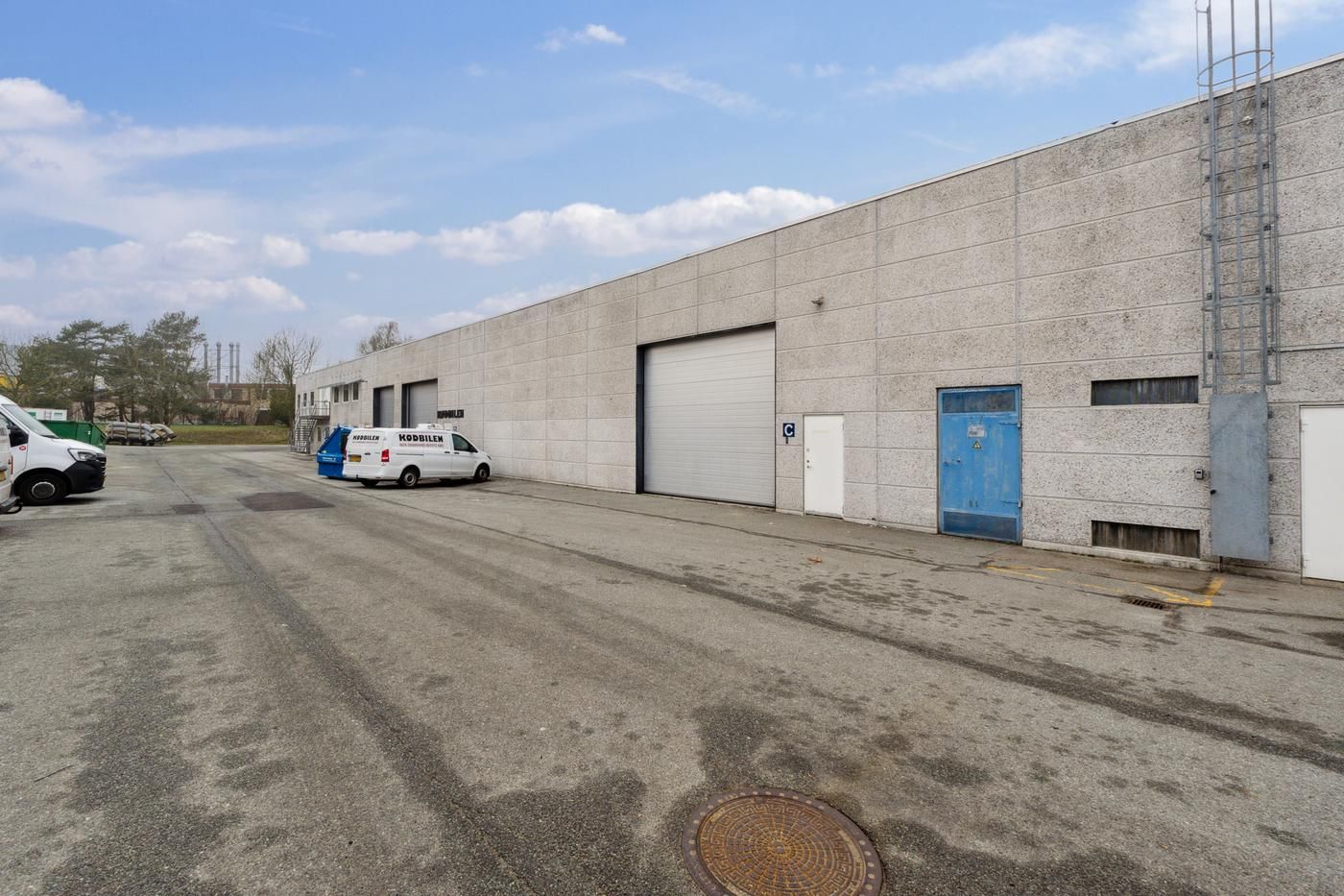 Lagerbygning med to store garageporte og en hvid dør samt en lyseblå dør, hvor to lastvogne er parkeret