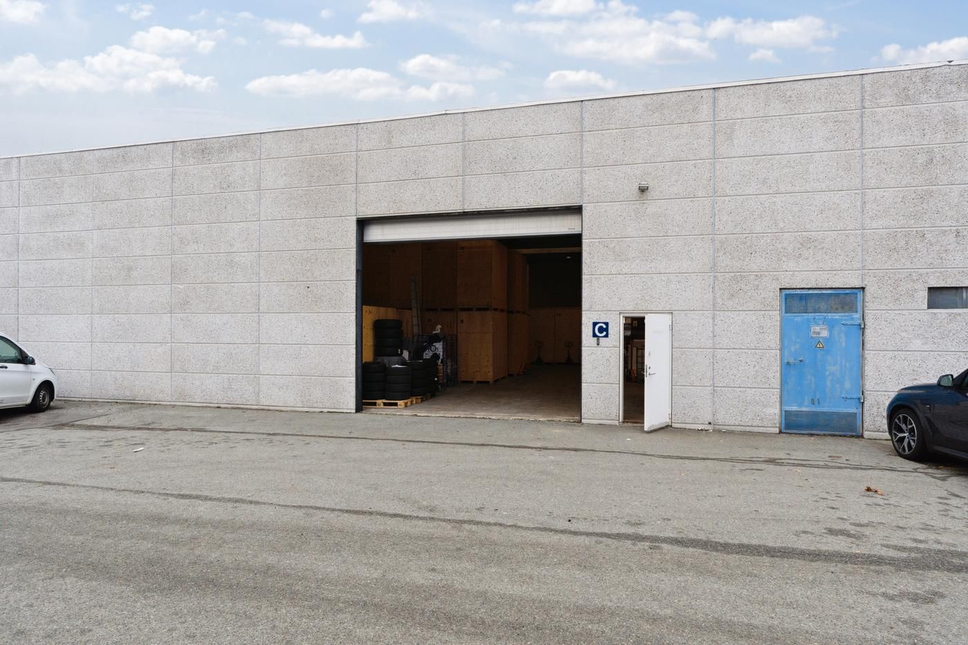 Lagerbygning med to garager og åben dør til parkeringsplads og lager med tømmerkasser og dæk