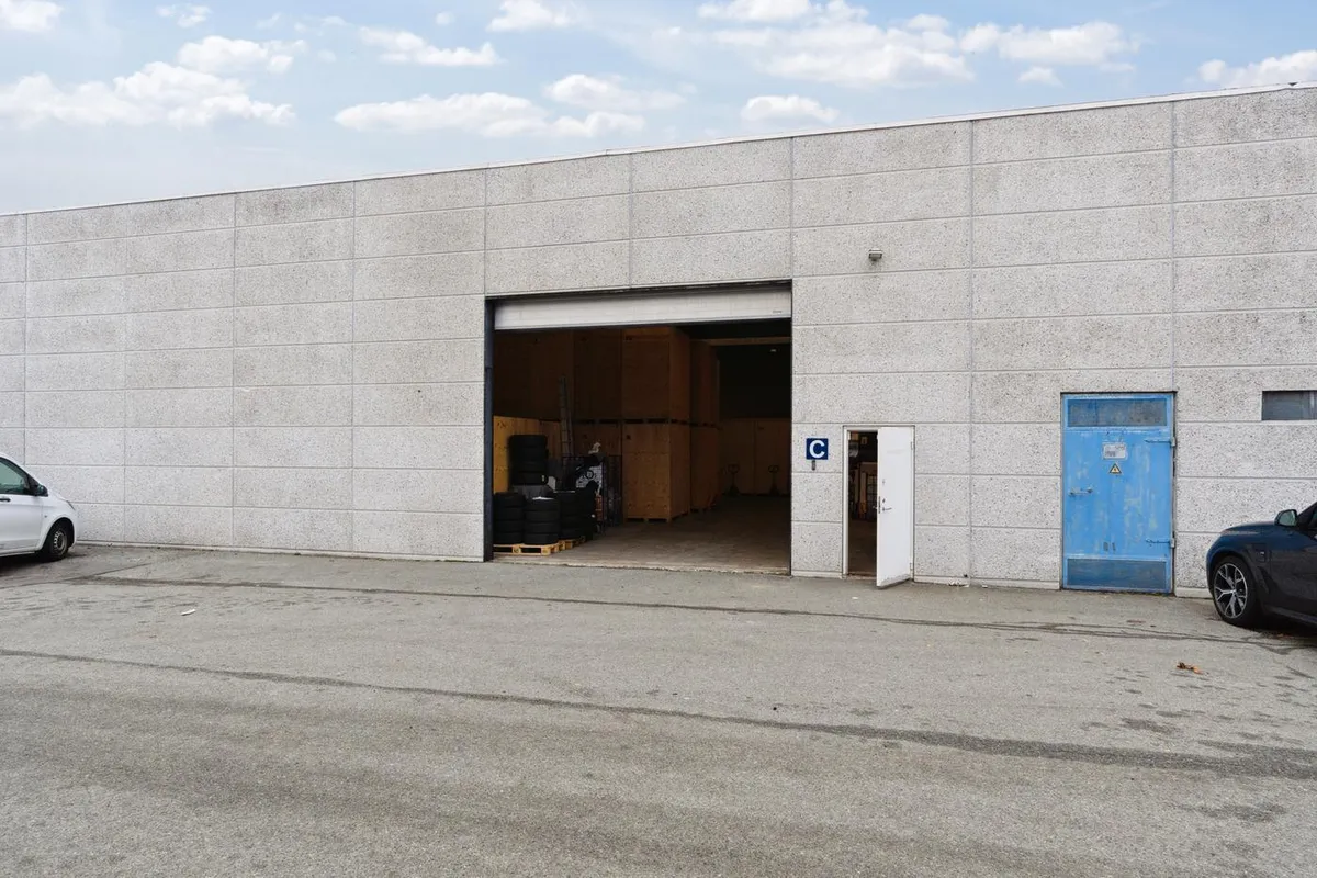 Lagerbygning med to garager og åben dør til parkeringsplads og lager med tømmerkasser og dæk