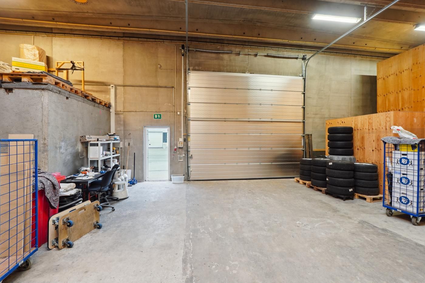 En stor lagerhal med to store porte, hvoraf den ene er åben, og indvendig er der en række dæk samt andre genstande.