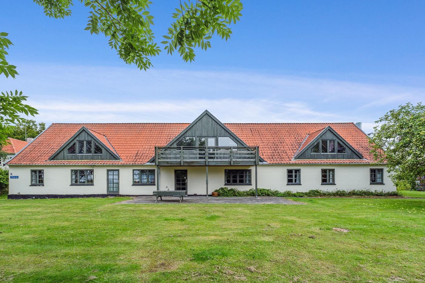 Hvidt hus med tegltag og balkon, have med græs og træer, vej, sol og skyer, parkeringsplads med bænk og grønne planter