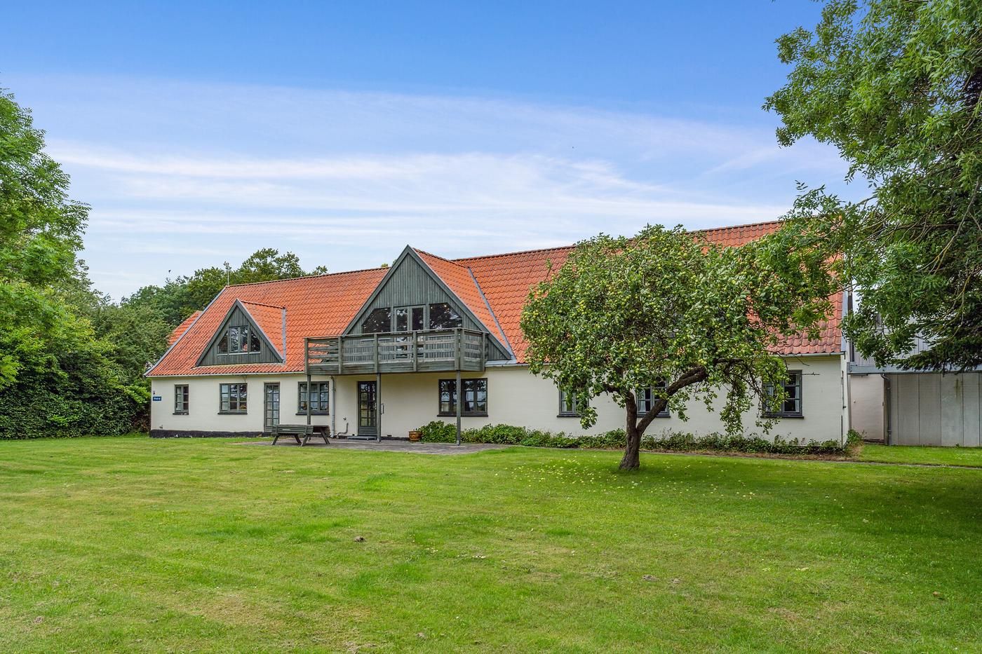 Hvidt hus med rødt tag og balkon og grøn græsplæne med et træ og blomster.