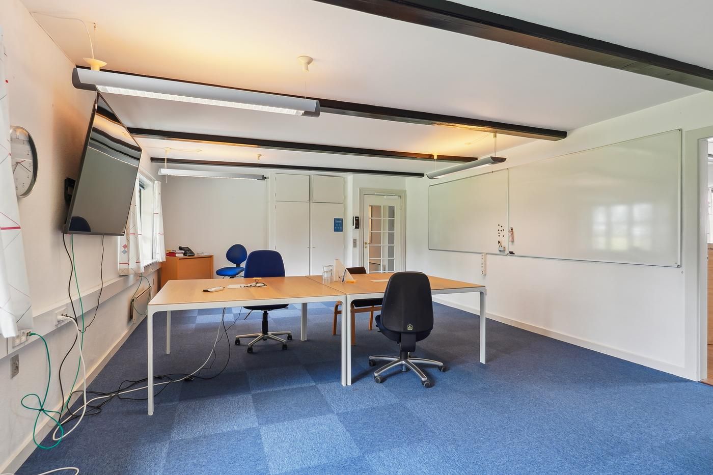 Kontor med skriveborde, stole og tv-skærm og tavle i lyserum med lyse gulve og loft.