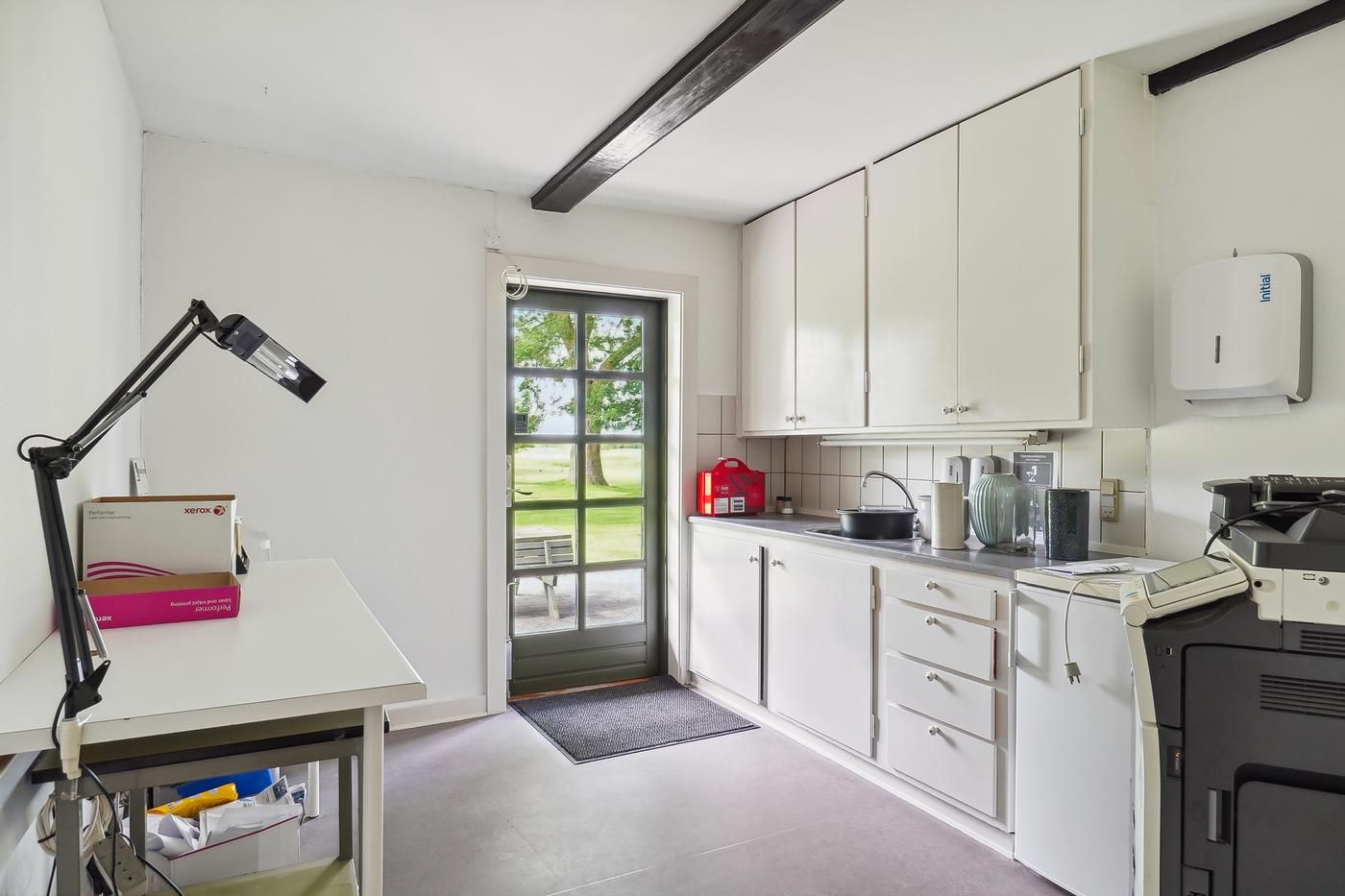Kontor med køkkenhjørne, skrivebord, bøsser og skrivemaskine samt vinduer med udsigt til park og græsplads