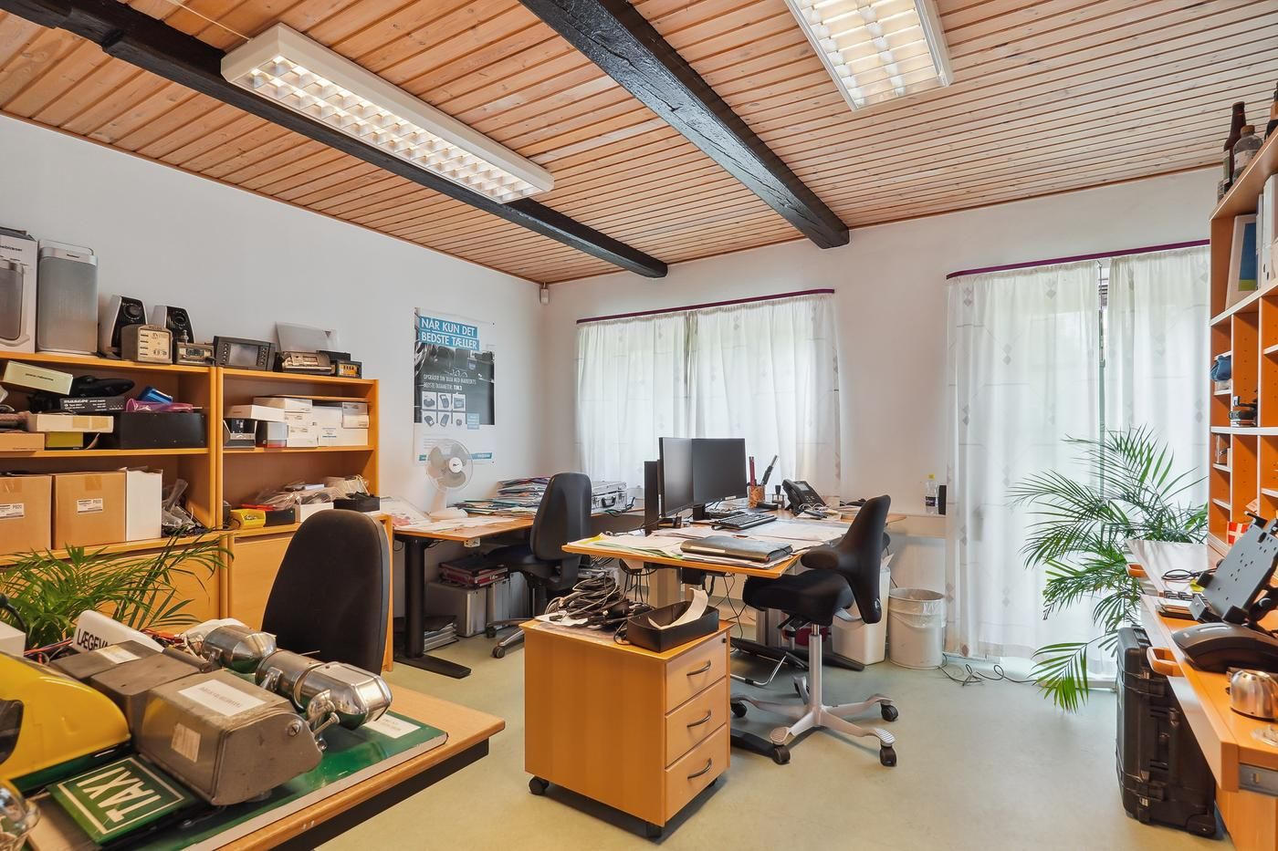 Kontor med to skriveborde, hver med computer og stol. Højt stativ med aviser og en ventilator. Væg med plakat og