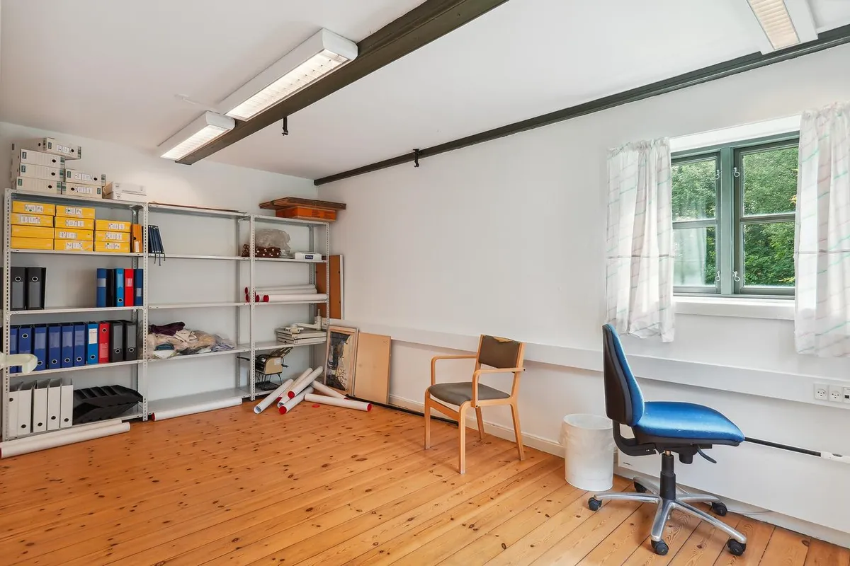 Et stort, tomt kontor med høje hvide vægge, trægulv og højtophængte lysekroner. Der er flere hylder med papirvarer og en