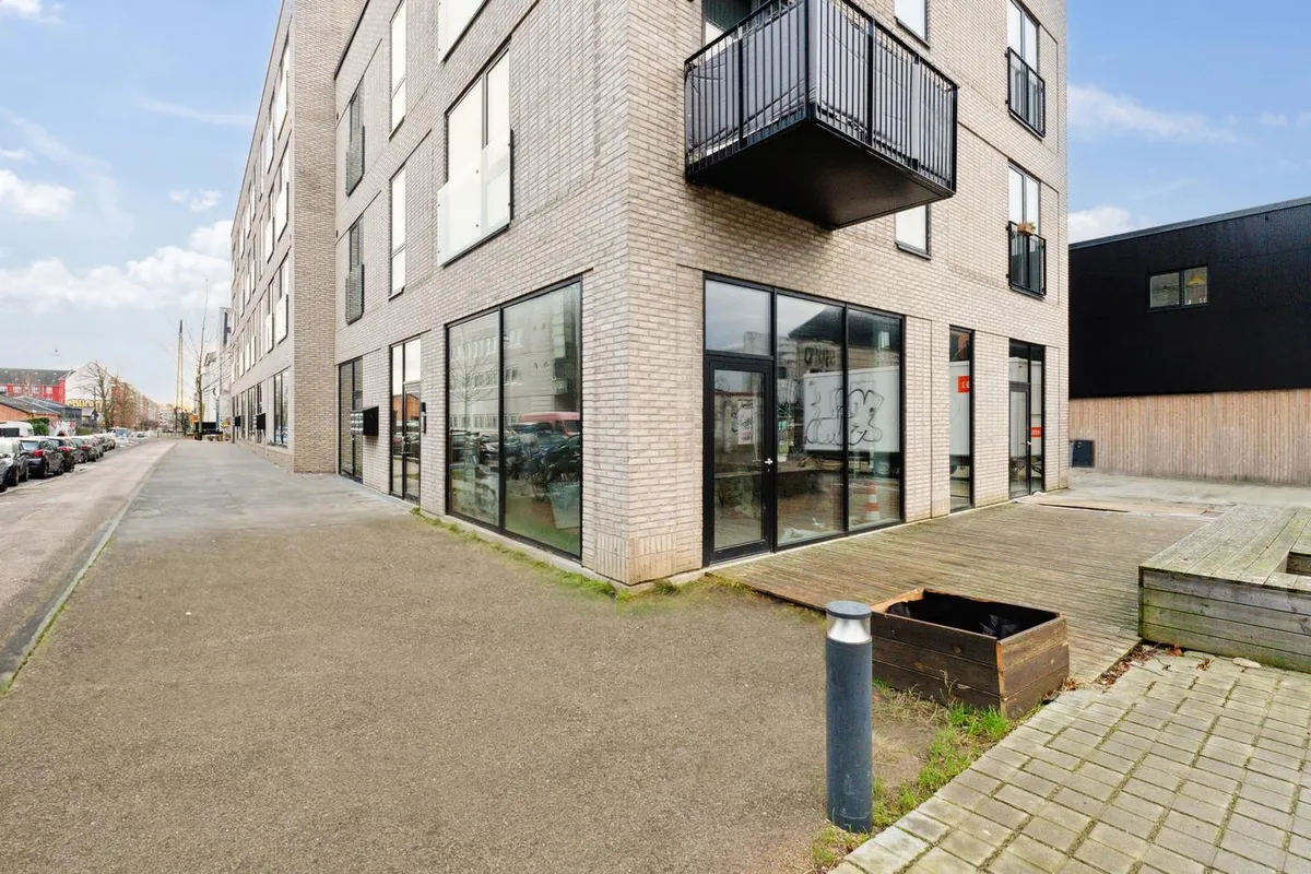 Hjørnelejemål med store vinduer og terrasse placeret i en bydel med mange parkerede biler og en bygning med sort facade