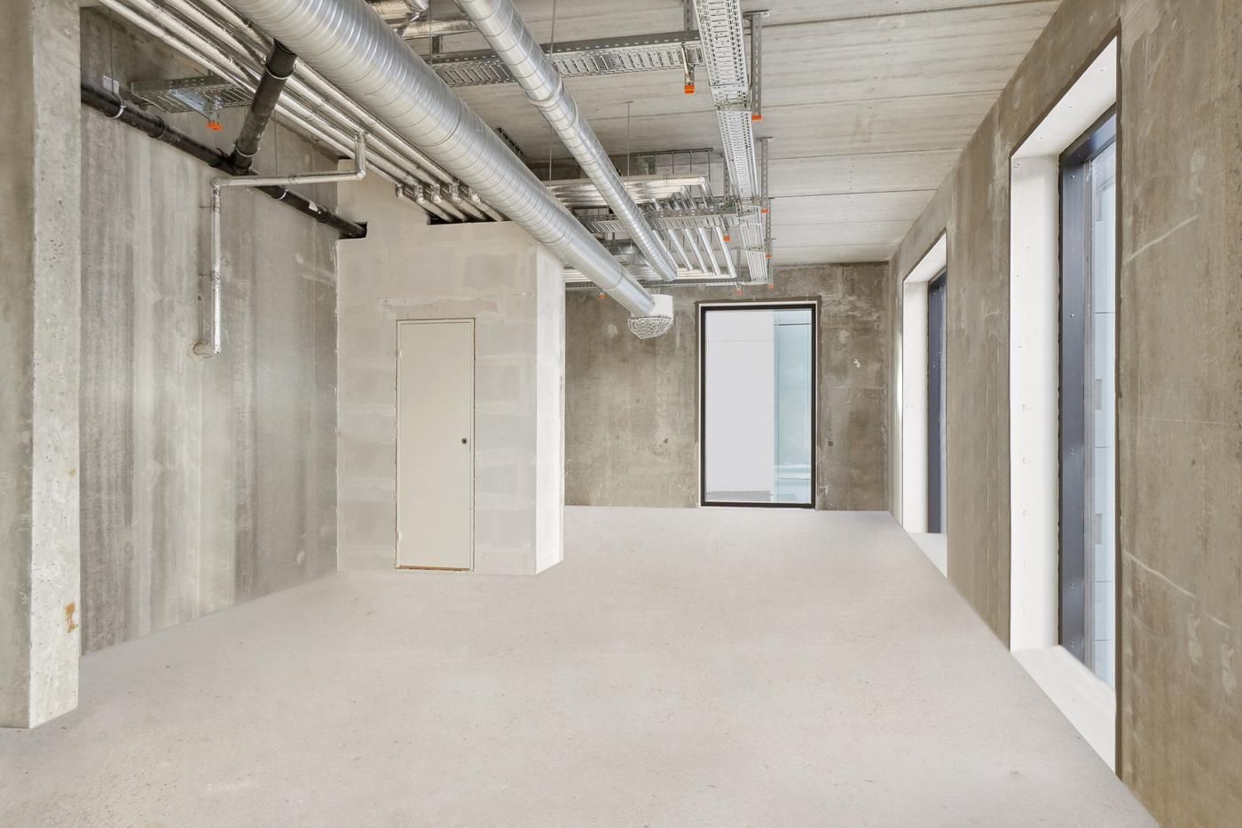 En stor, tom rum med hvide gulv og høje, lyse vægge. Der er tre vinduer og et hvidt dørblad i væggen til venstre.