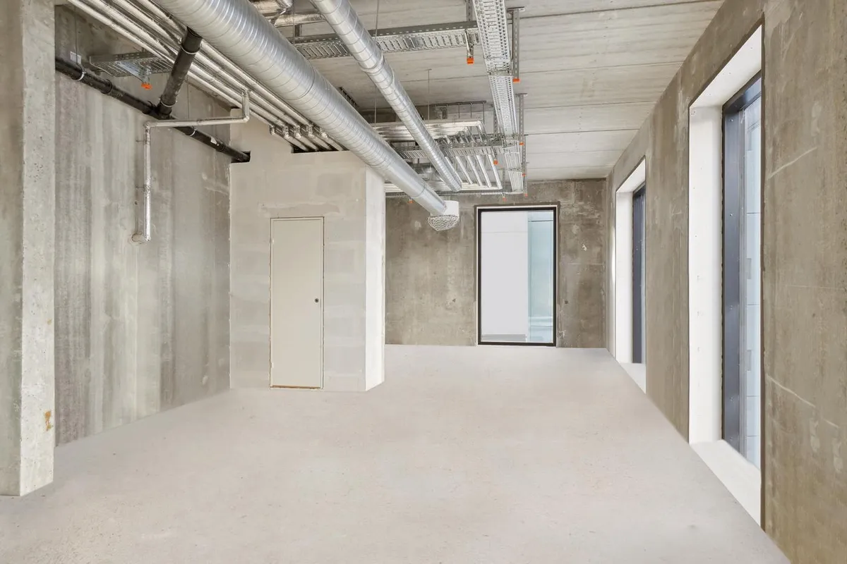 En stor, tom rum med hvide gulv og høje, lyse vægge. Der er tre vinduer og et hvidt dørblad i væggen til venstre.