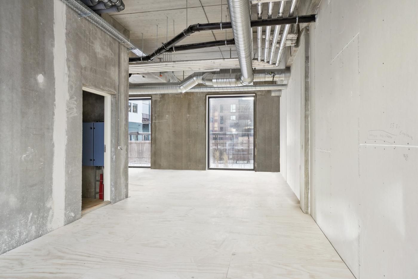 Kort gange med lyse gulve og hvide vægge og vinduer med udsigt til gårdhave. Væggene er af beton og der er røre og slanger