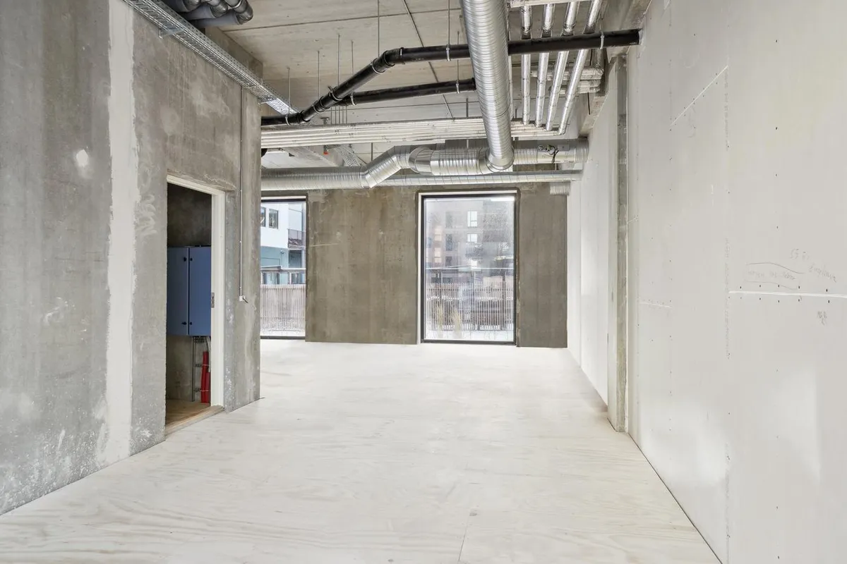 Kort gange med lyse gulve og hvide vægge og vinduer med udsigt til gårdhave. Væggene er af beton og der er røre og slanger