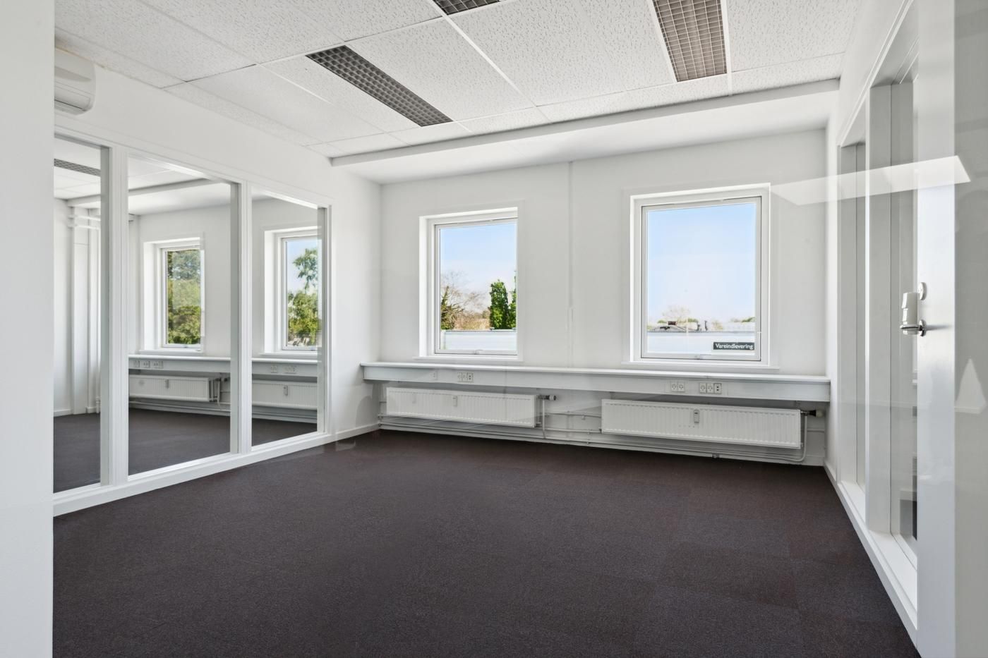 Foto af et tomt kontor med hvide vægge og gulv og vinduer med udsigt til grønne træer og lyse himmel