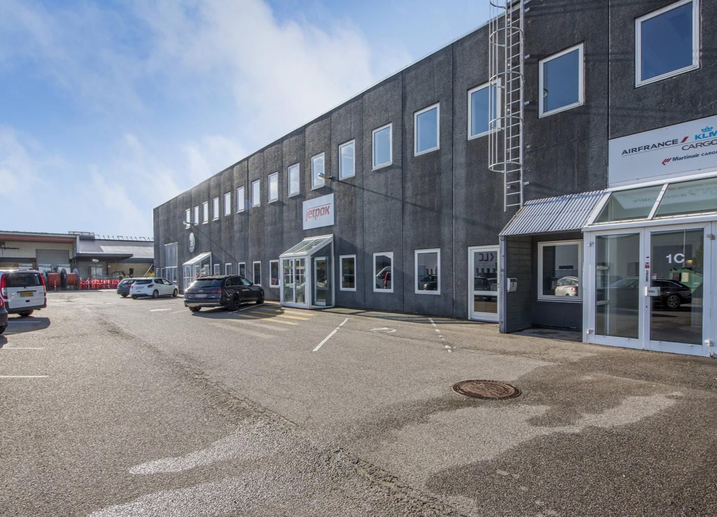 Moderne kontorbygning med Air France Cargo og Jetpak skilte samt parkeringsplads med parkerede biler og spor på asfalten