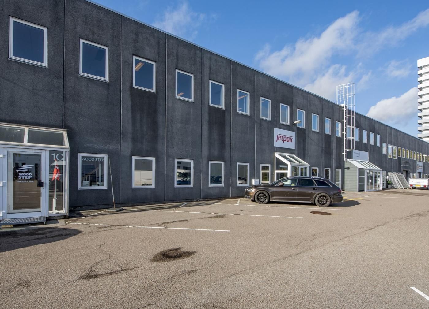 En parkeringsplads med en parkeret sorte Audi A6 foran en Jetpak kontorbygning med glasruder og glasdøre