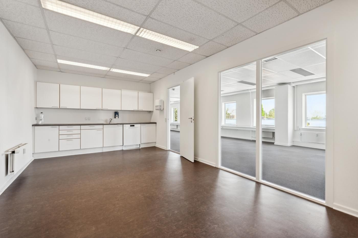 Et nyt kontor med hvide vægge og lysegult gulv, to enheder med store glasruder, hvide køkkenskabe og moderne lys.