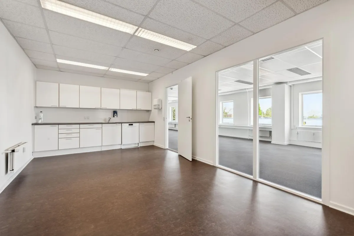 Et nyt kontor med hvide vægge og lysegult gulv, to enheder med store glasruder, hvide køkkenskabe og moderne lys.