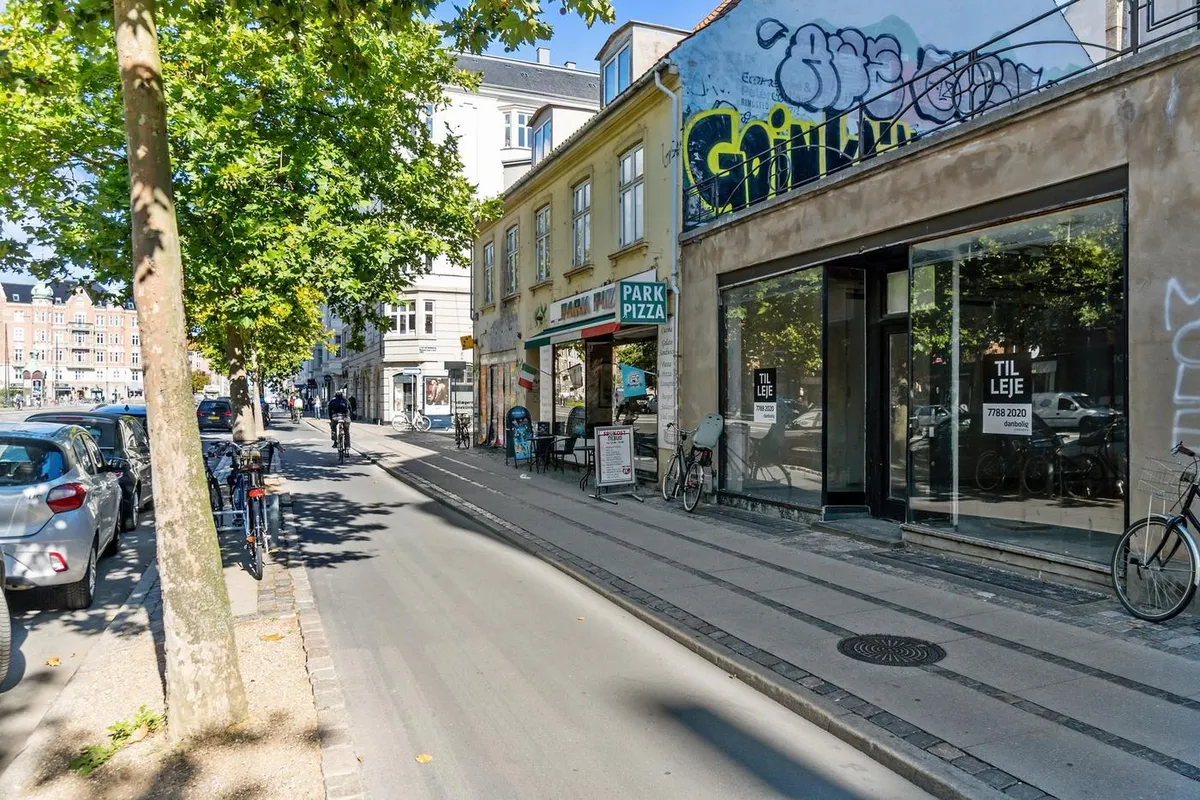 Østerbrogade med parkerede biler, cykler, grønne træer og bygninger med graffiti og butikker som Park Pizza