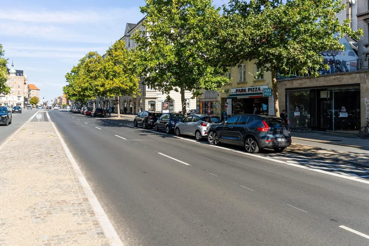 Østerbrogade med parkerede biler og en pizzeria med graffiti. Mange bygninger med store vinduer og grønne træer på fortovet.