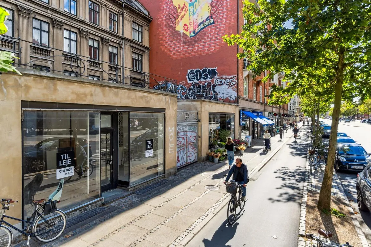 Man kører på cykel på Østerbrogade, hvor der er flere butikker og træer langs gaden