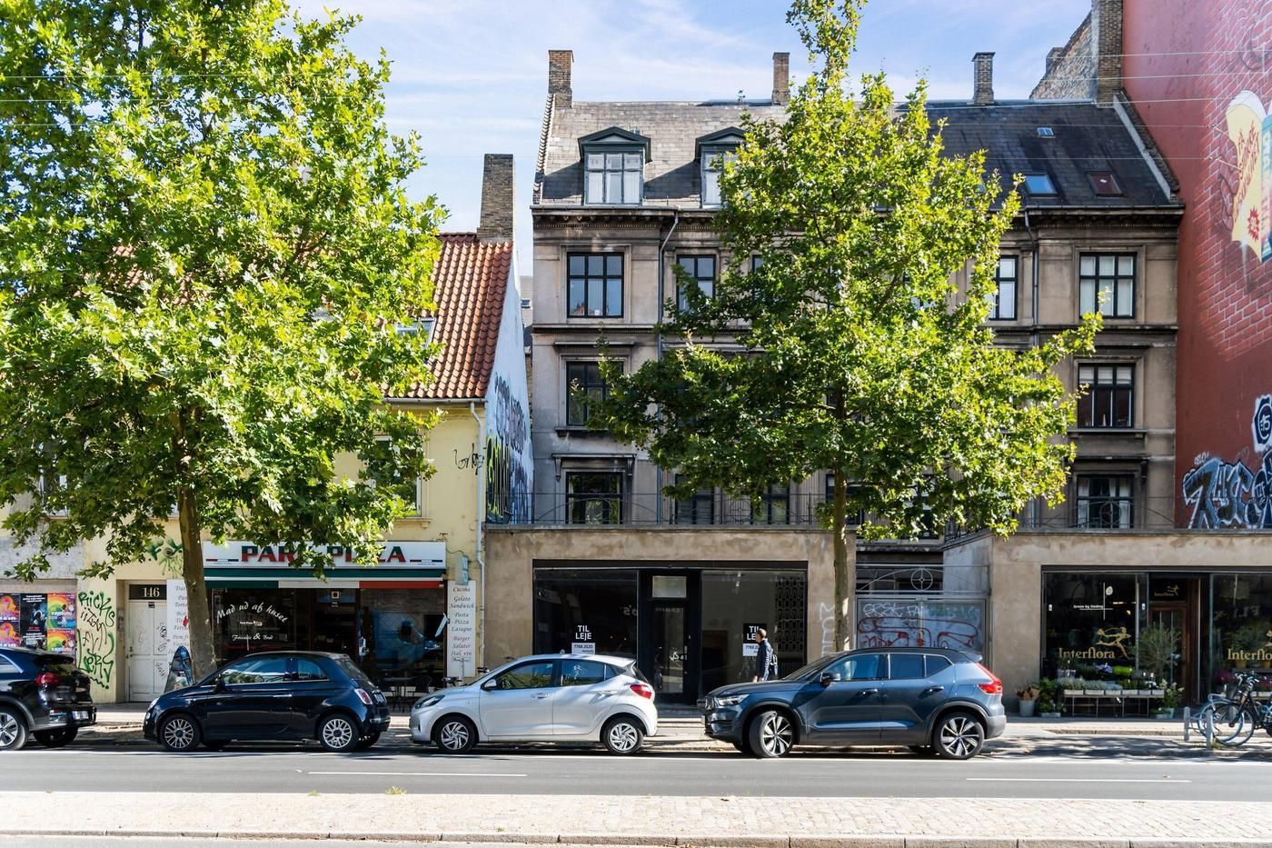 Erhvervslejemål med store vinduer på Østerbrogade med tre parkerede biler og en person med en rygsæk