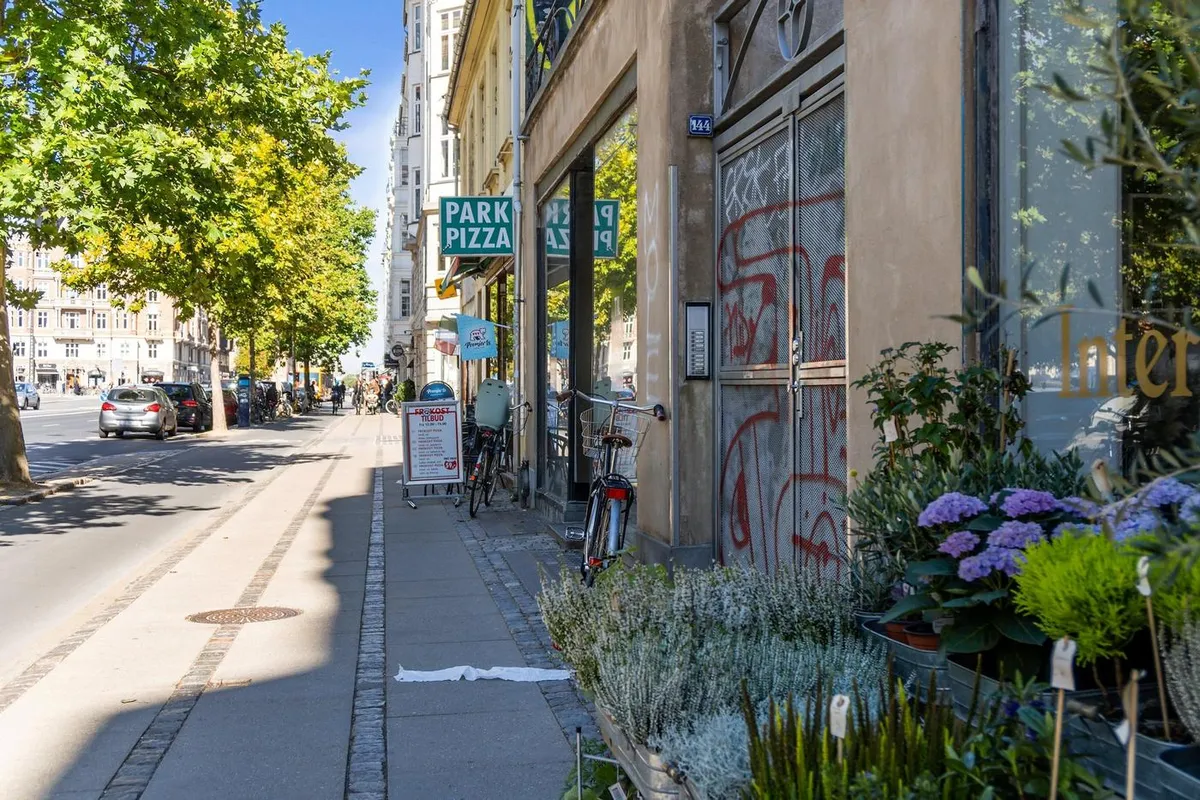 Parkerede cykler udenfor en pizzarestaurant, blomster i kasser og graffiti på facaden, samt biler og folk på gågaden