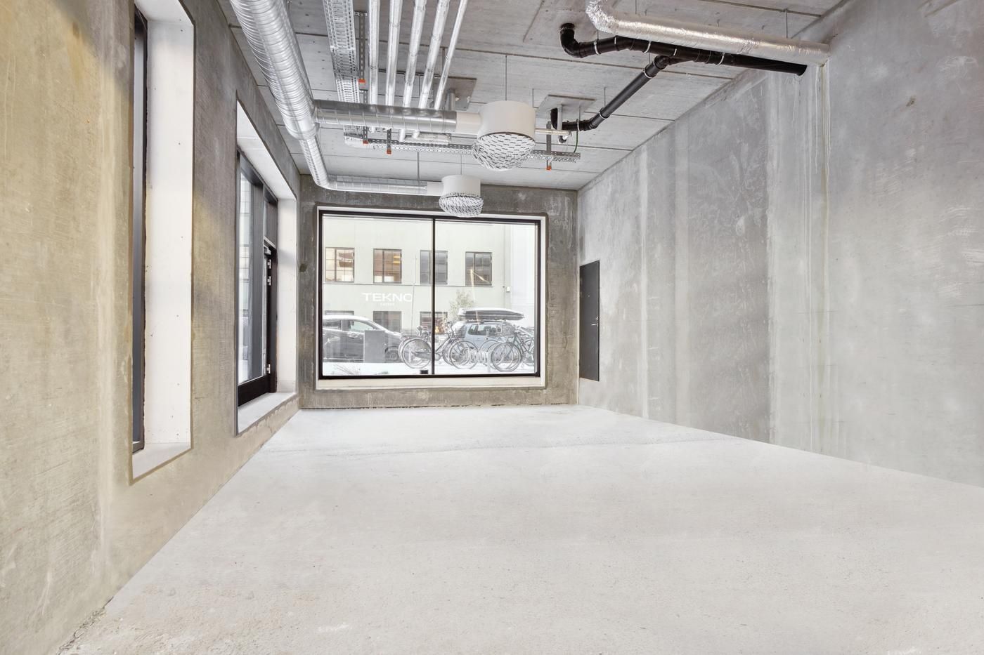 Rømmet rum med højt loft og metalrør, hvide vægge og vinduer med udsigt til parkerede biler og cykler udenfor