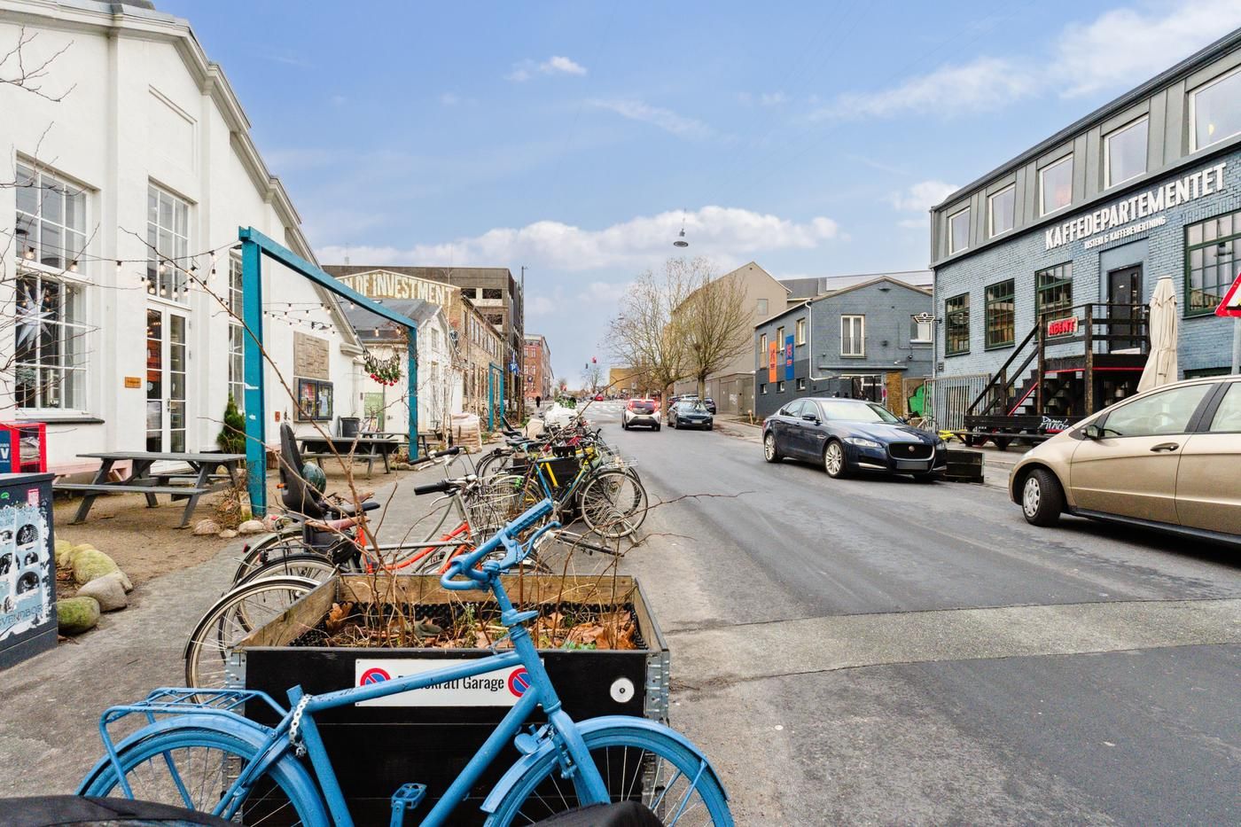 Kaffe og mad i nærheden af byens hovedvej. To biler står parkeret og to biler kører langs vejen. En bycykelparkering er på