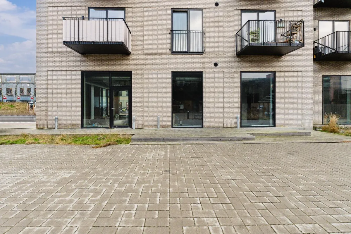 Bygningen med lyse vinduerne og terrasserne. Foran er en terrasse med stengulv. På hjørnet er der en skrånende grøn bakke