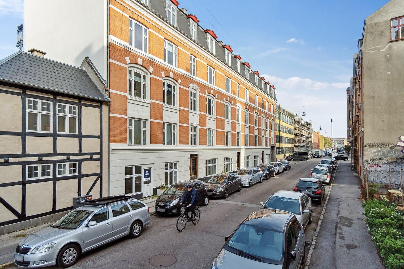 En cykelkører kører på en bred gade, der er omgivet af parkerede biler, forretningsbygninger og et hus med graffiti.