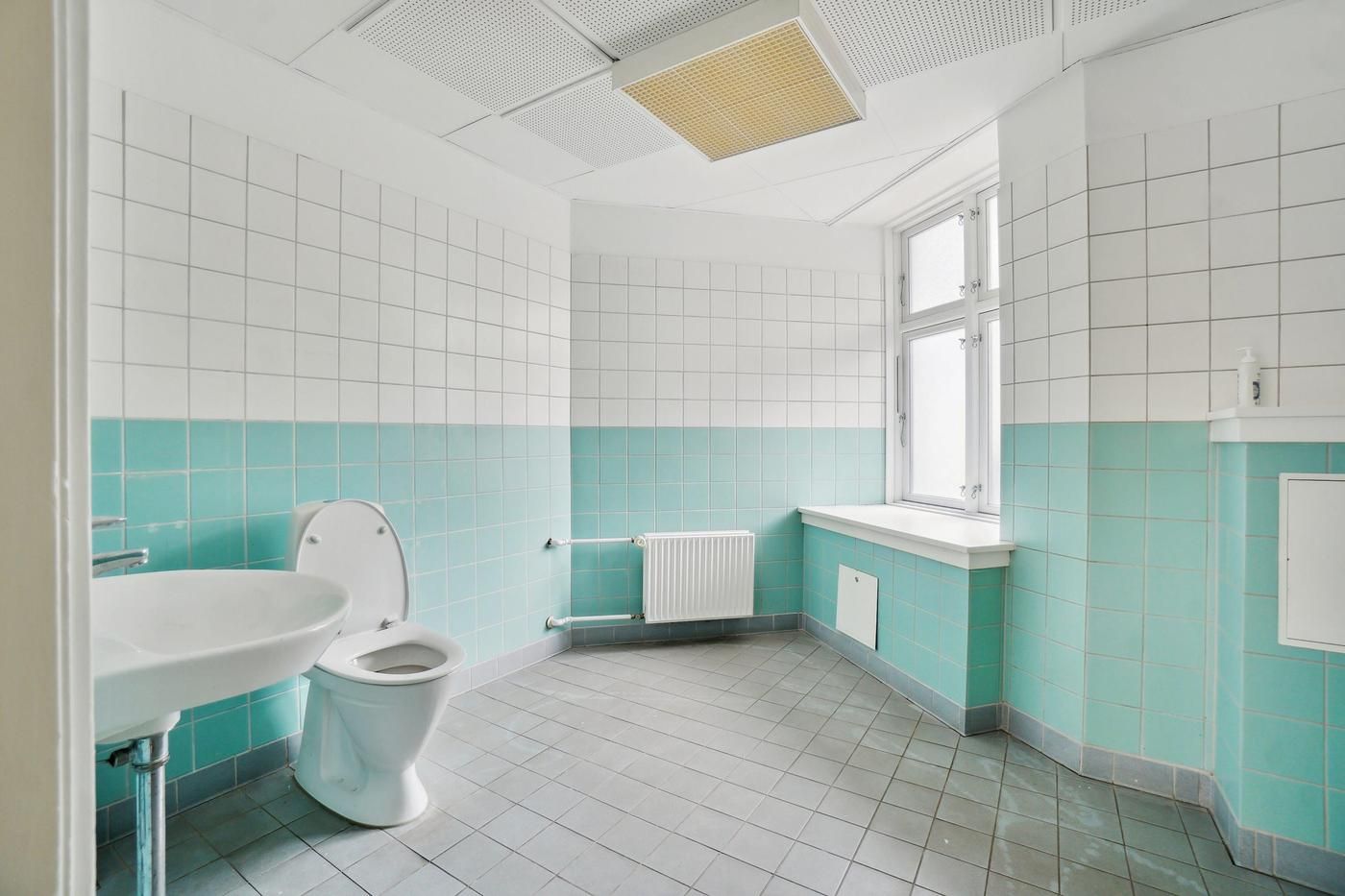 Et enkelt, lille badeværelse med en toilet, en hvid vask og en radiator i væggen. Hvid og blå kakelygter.