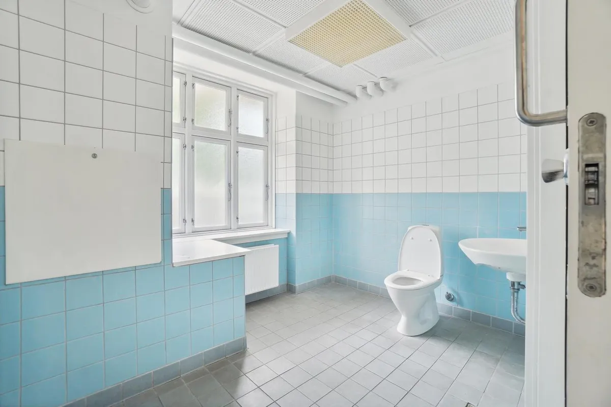 Badeværelse med toilet, hvidt vindue, lyseblå og hvide fliser og åben dør