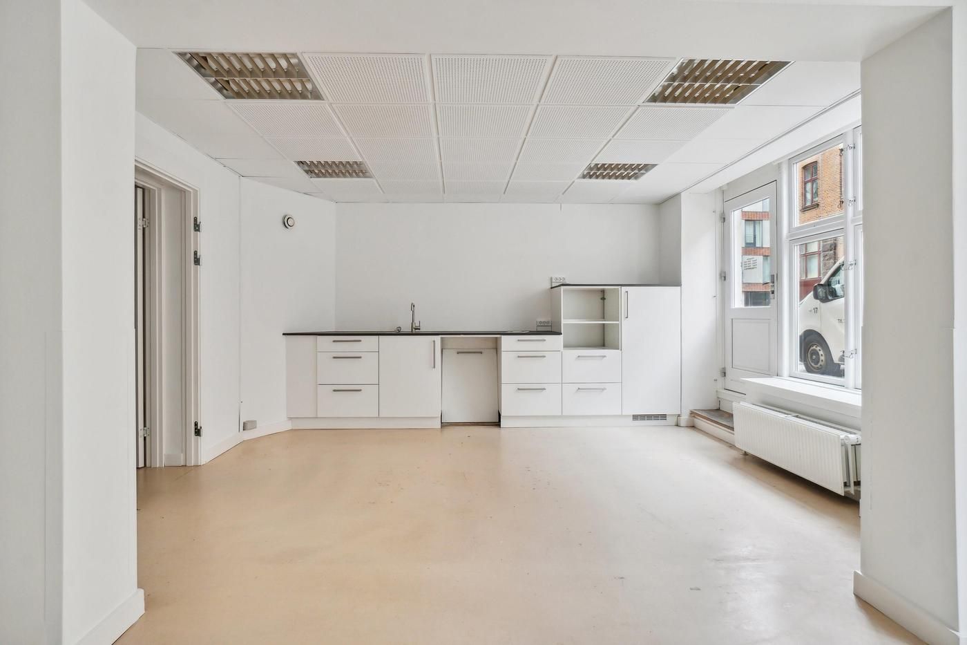Minimalistisk indrettet erhvervslejemål med hvide vægge og loft, køkken med hvide skabe og sink, og vinduer med udsigt til