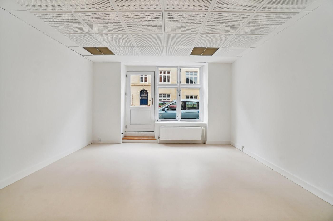 Hvid, tom stue med åbne døre og vinduer, hvor der ses biler og en bygning udenfor