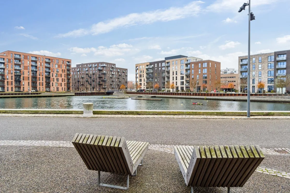 To træstole står ved en flod i en by med bygninger i baggrunden og en sø i midten