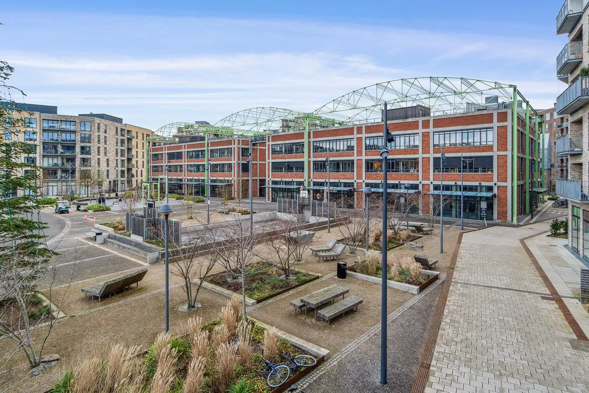 Anlægget med flere bygninger og grønne områder og parkerede biler under en blå himmel med skyer