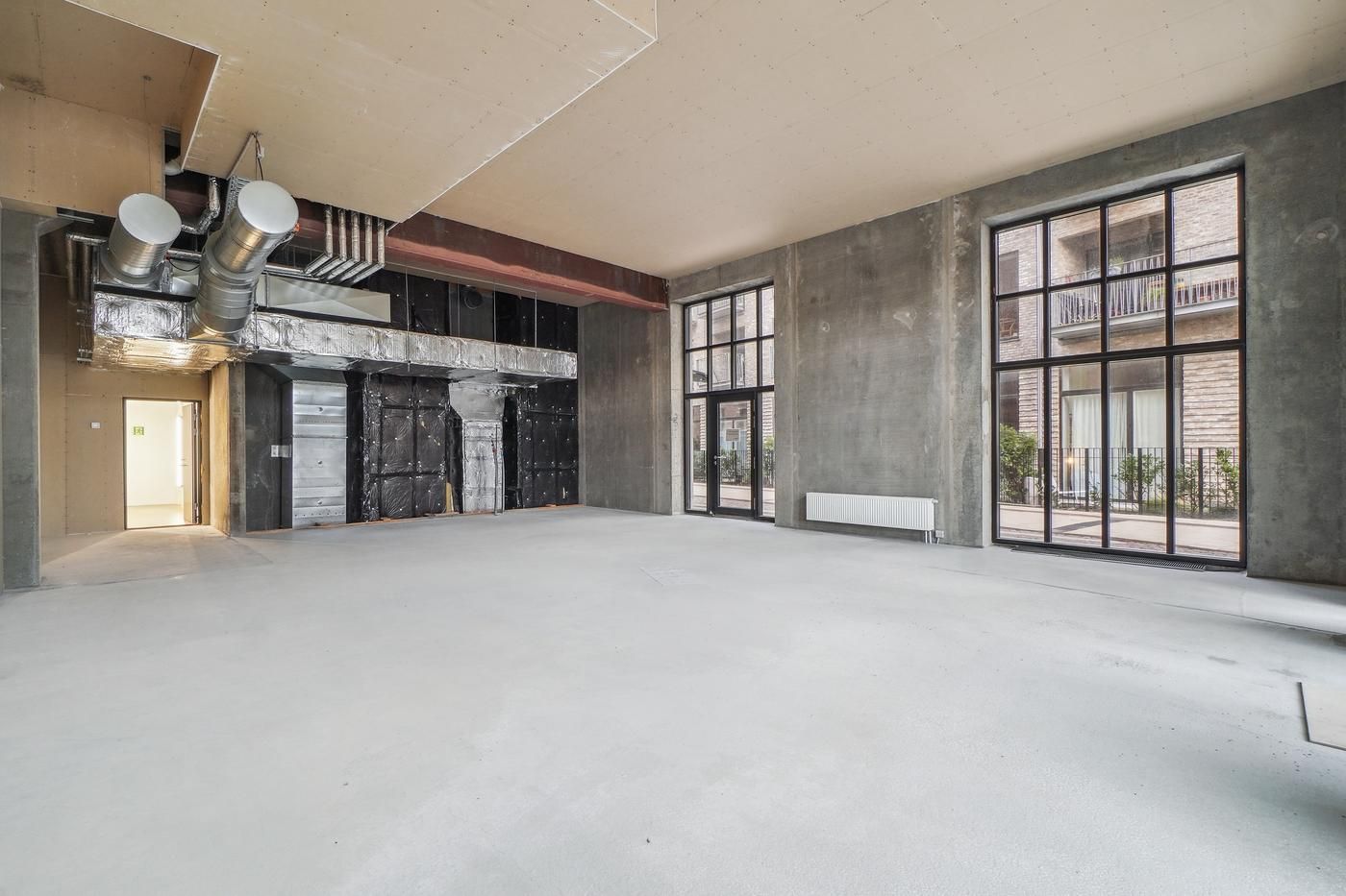 Et tomt rum med højloft og store vinduer, et hvidt gulv og en dør til en anden rum er synlig