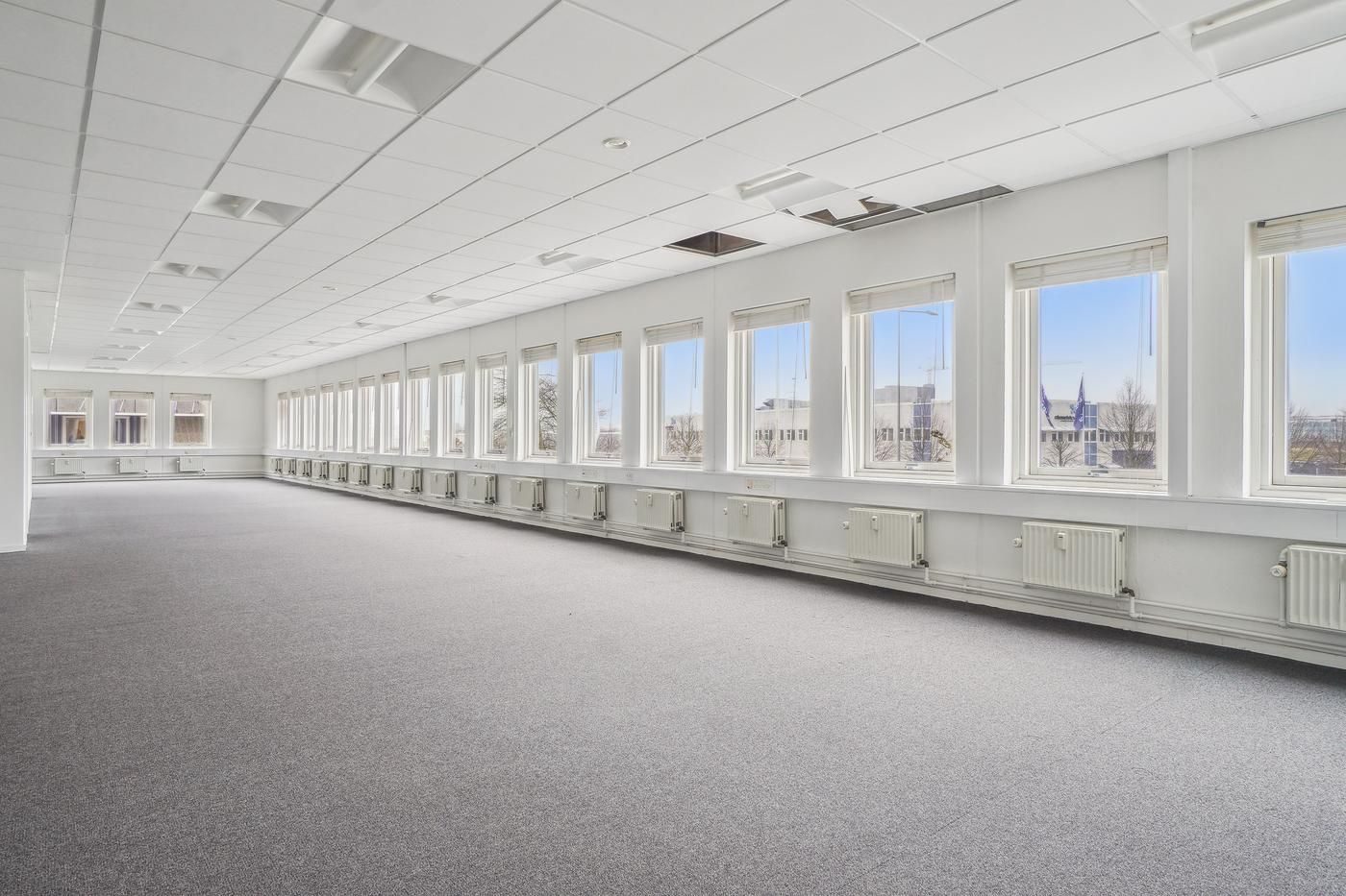 En stor lys og velholdt kontorrum med glasvinduer og gulvbelægning i lysegråt og hvide vægge med radiatorer og luftkanaler