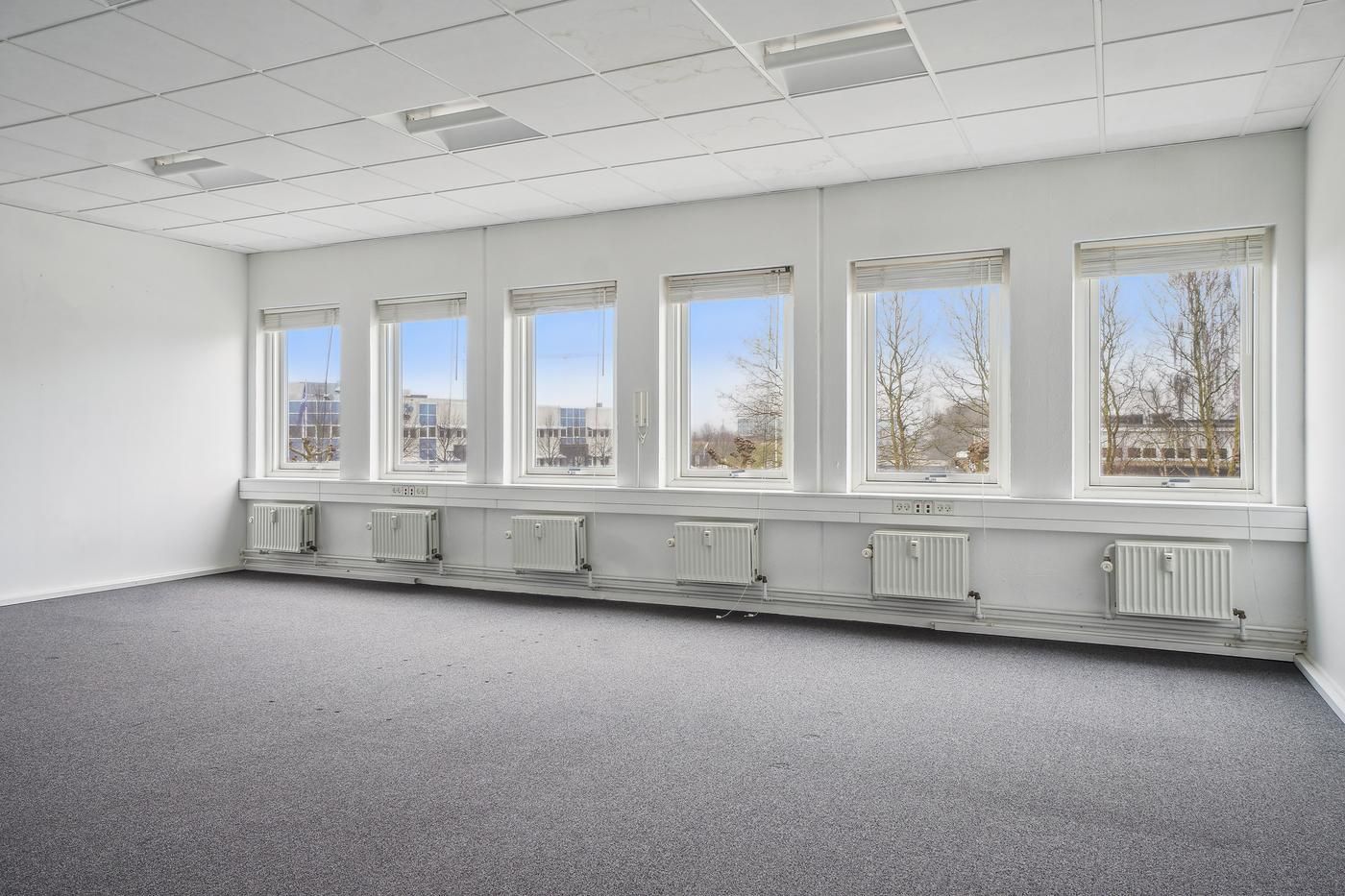 Rummelige kontorlokaler med hvide vægge, gulvbelægning, radiatorer og vinduer med gardiner samt udsigt til bygninger og træer