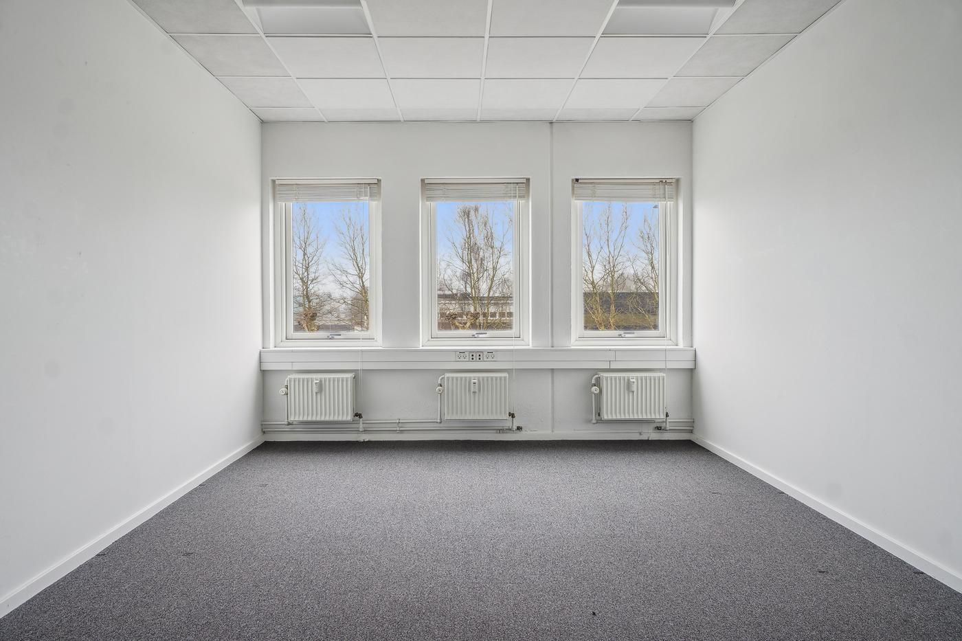 Lukket kontor med tre vinduer og hvide vægge, gulv og loft samt tre radiatorer under vinduet og stikkontakter i væggen