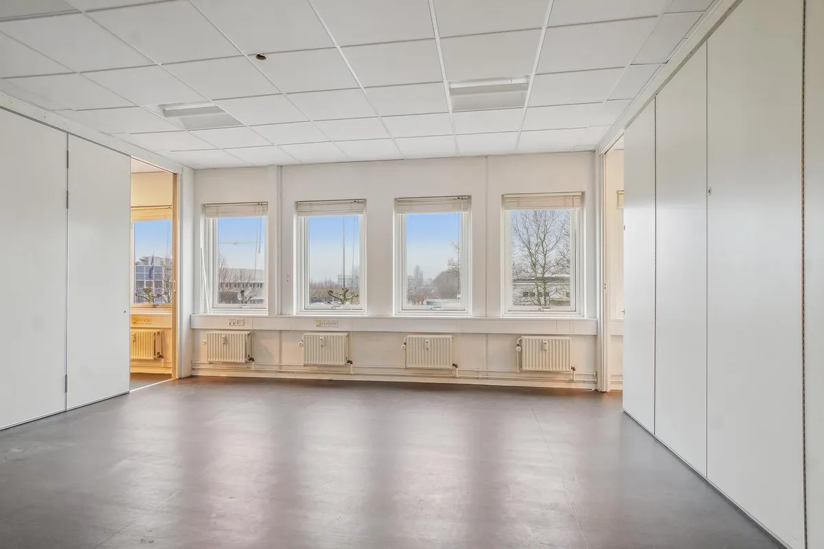 En lys og ren kontorrum med hvide vægge, gulv og loft samt fire store vinduer og flere radiatorer under hvert vindue.