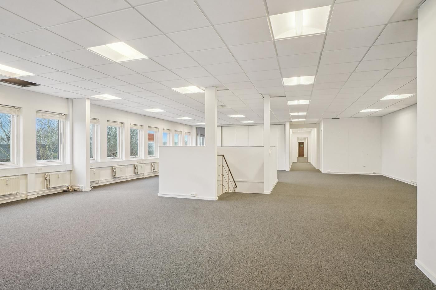 Et tomt kontor med hvide vægge, lyse gulvbelægning og store vinduer med udsigt til træer udenfor