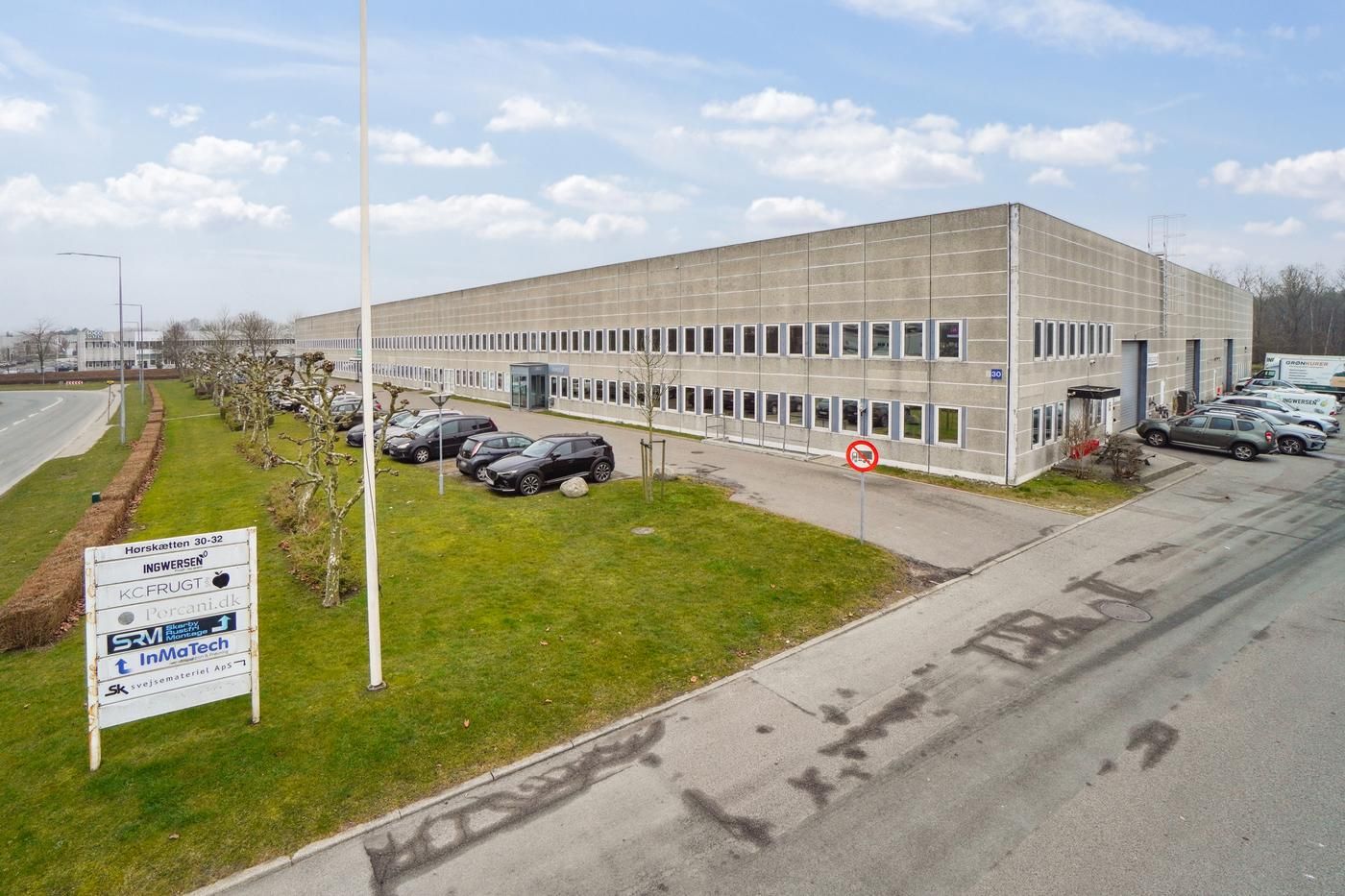 Et stort kontorbyggeri med parkeringsplads og græsplæne med forskellige firmaer og biler parkeret udenfor.