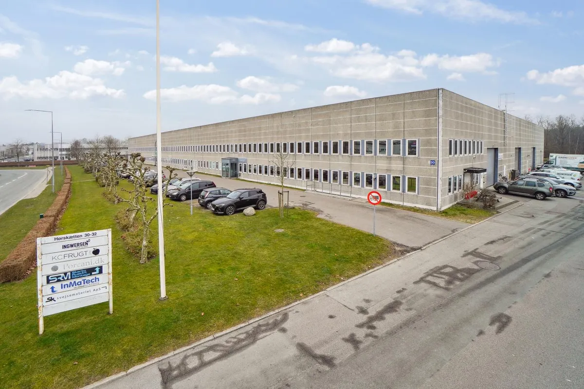 Et stort kontorbyggeri med parkeringsplads og græsplæne med forskellige firmaer og biler parkeret udenfor.