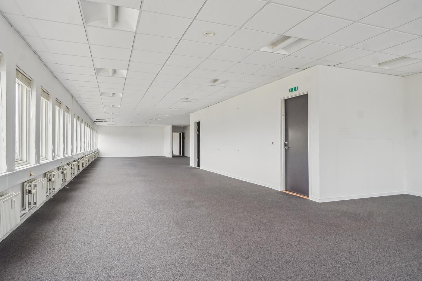 Et stort og lyst kontor med hvide vægge og grå gulv. I loftet er der en hvid indretning med flere lampetyper. Et af rummene