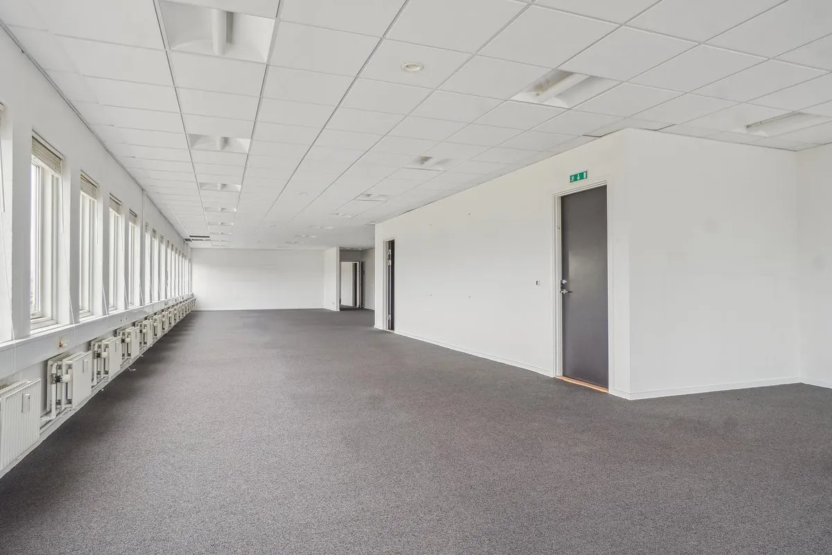 Et stort og lyst kontor med hvide vægge og grå gulv. I loftet er der en hvid indretning med flere lampetyper. Et af rummene