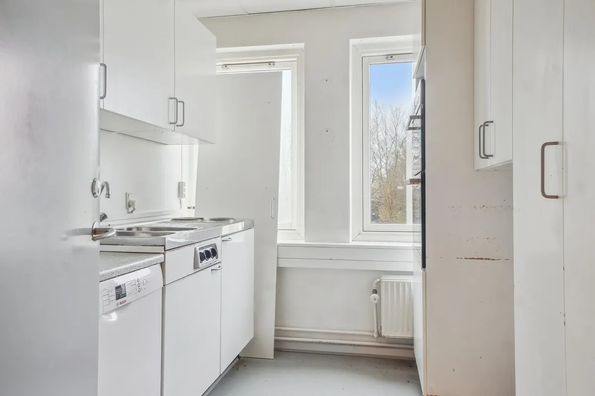 Køkken med hvide vægge og møbler, spisestue med lysevinduer, spisestue med radiator og dør, hvid ovn og tekøkken med en